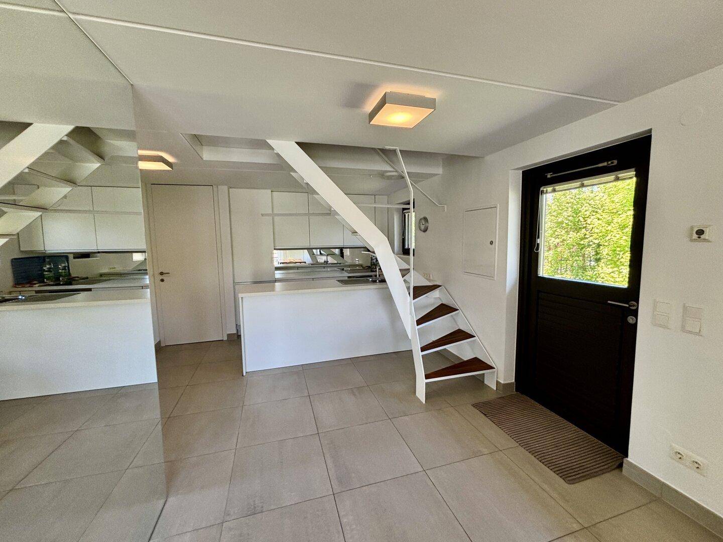 Eingangsbereich mit moderner Treppe, Fliesenboden und Blick auf die offene Küche.