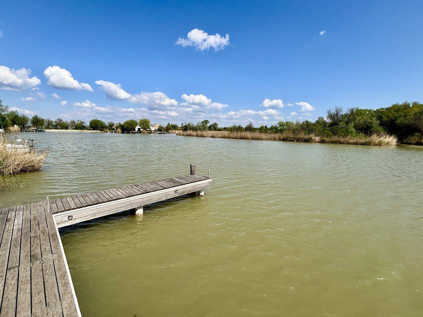 Malerischer Blick auf den See mit einem Holzsteg unter blauem Himmel und weißen Wolken.