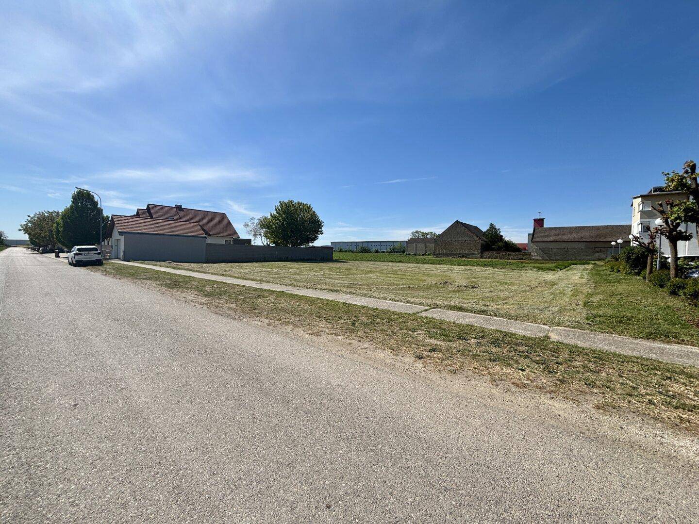 Offenes Grundstück mit Straße und Gehweg im Vordergrund, umgeben von ländlicher Bebauung und Natur.