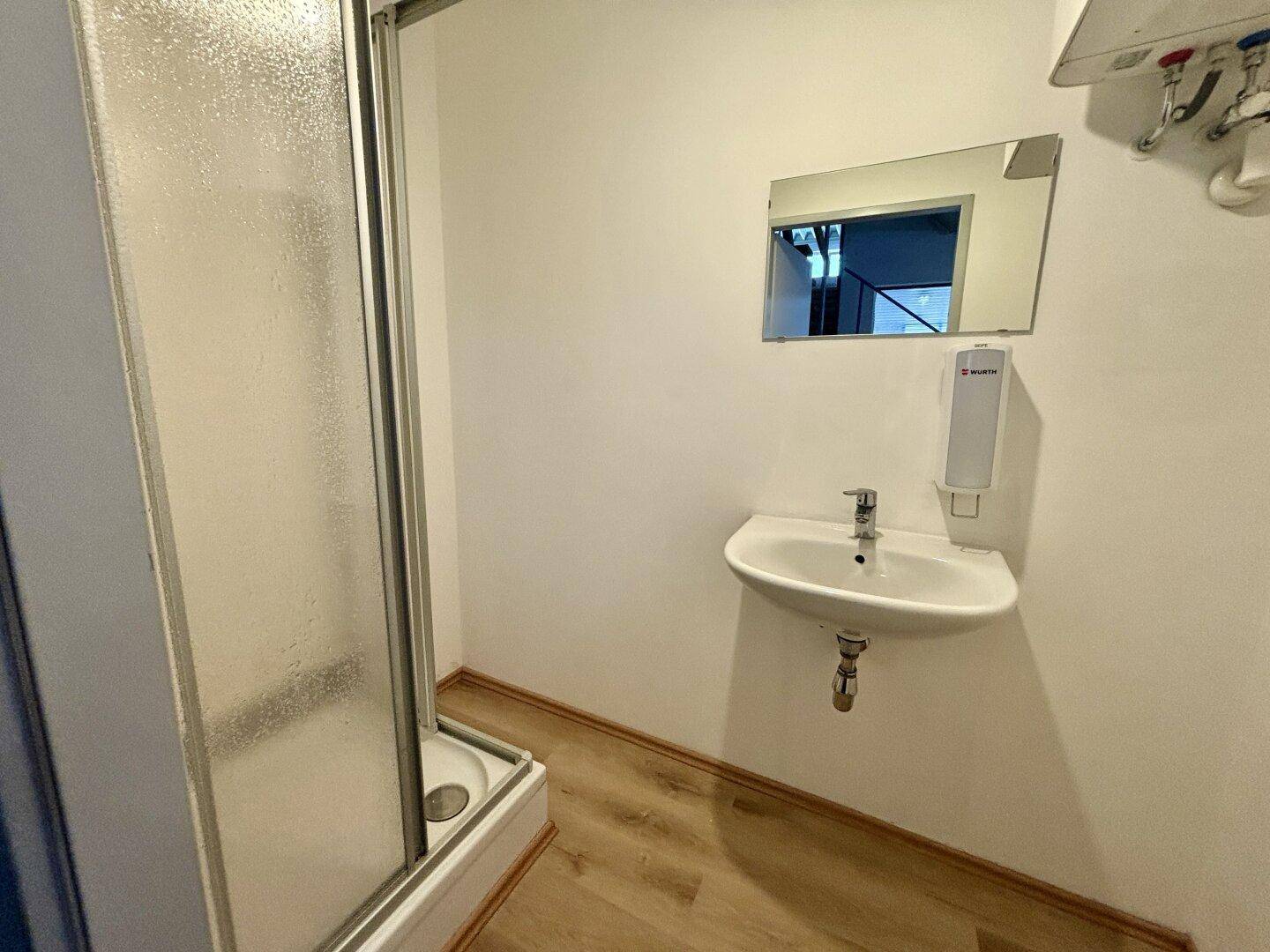Kleines Badezimmer mit Duschkabine, Waschbecken und Spender an der Wand.