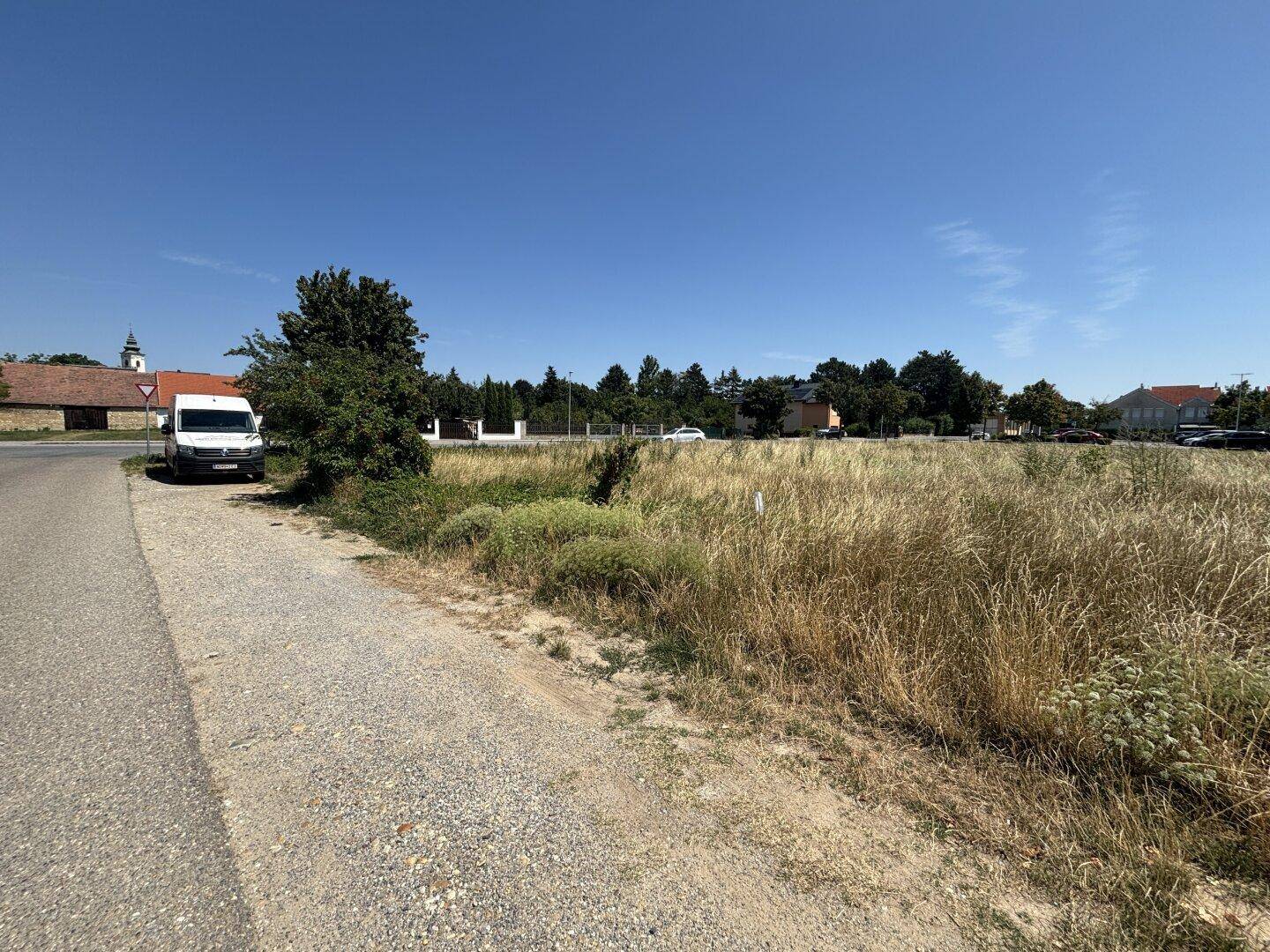 Unbebautes Grundstück mit Schotterweg und Blick auf eine Kirche sowie weitere Gebäude in der Ferne.