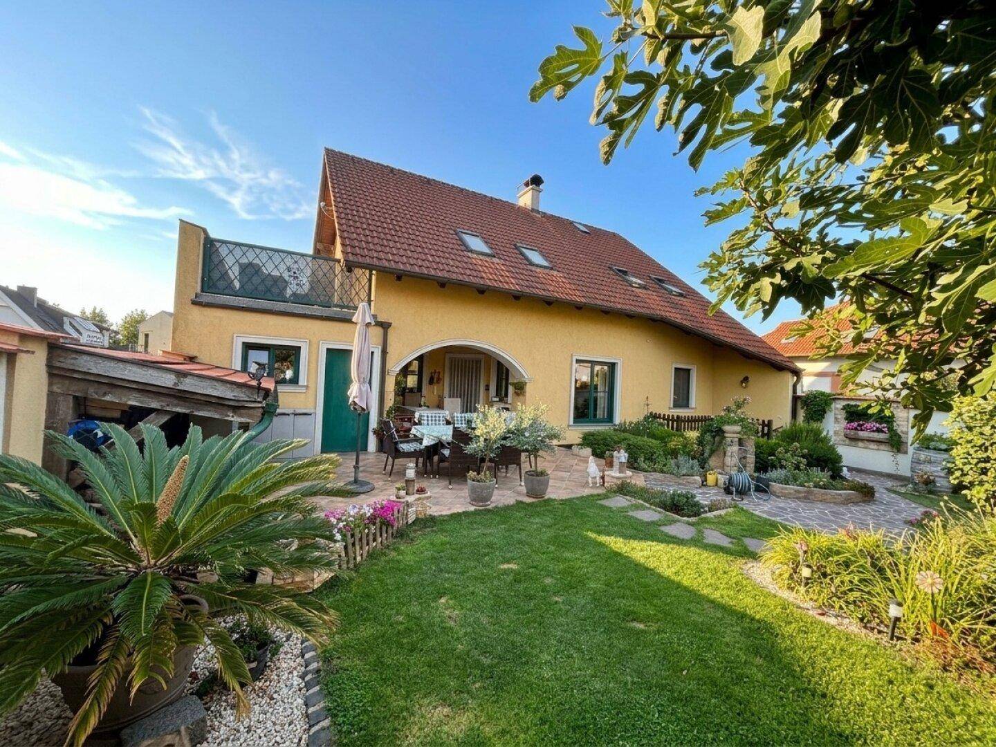 Rückansicht des Hauses mit rotem Ziegeldach, gepflegtem Garten und überdachtem Sitzbereich.