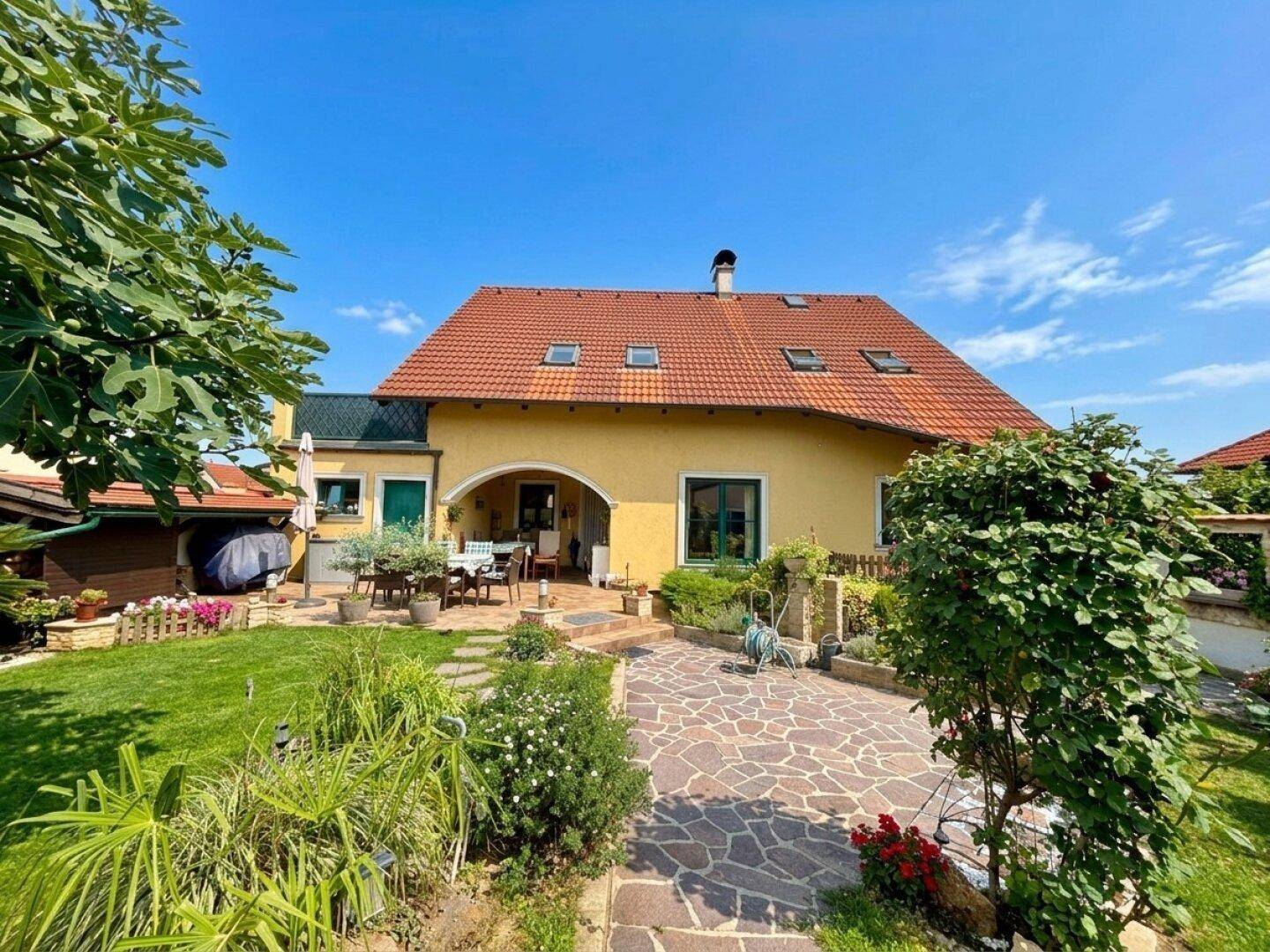 Rückansicht des Hauses mit rotem Ziegeldach, gepflegtem Garten und überdachtem Sitzbereich.