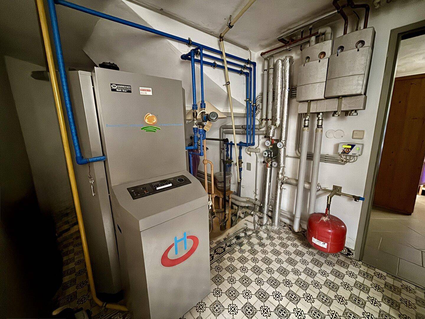 Heizungsraum mit großem Heizkessel, Rohrleitungen und gemustertem Fliesenboden.