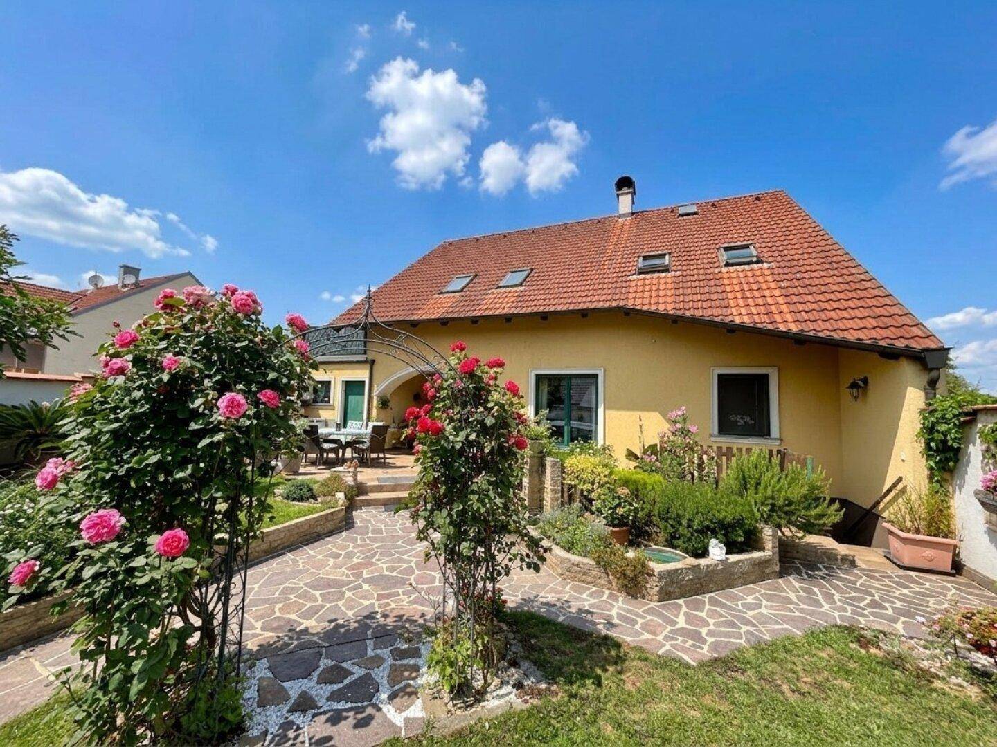 Hausfassade mit rotem Ziegeldach, umgeben von blühenden Rosensträuchern und einem gepflasterten Weg.