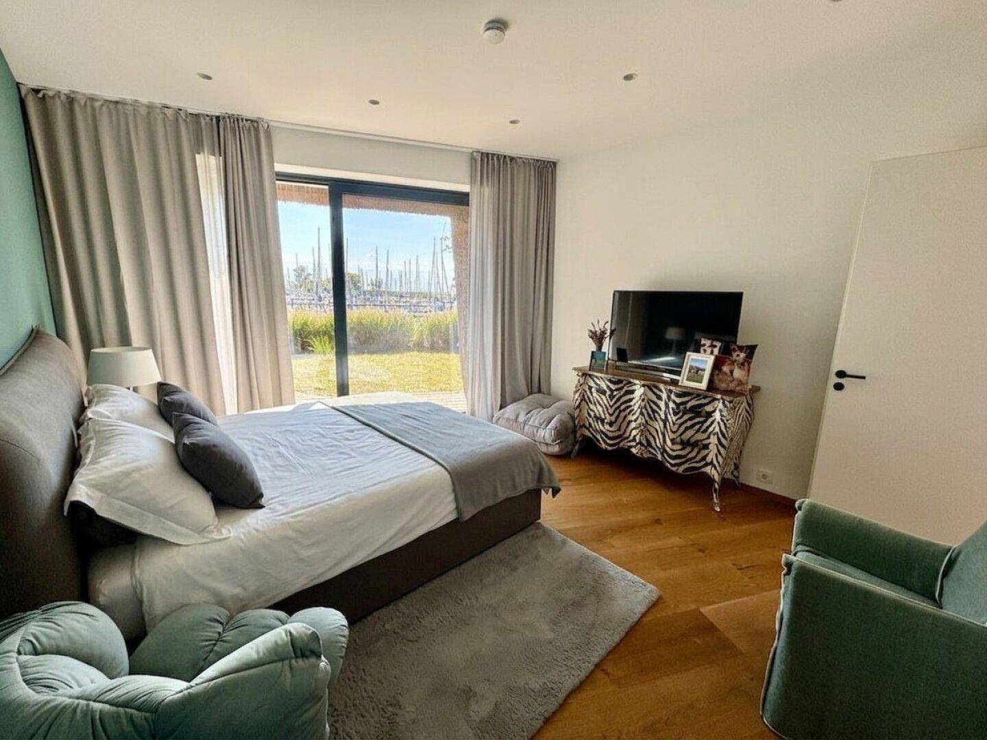 Helles Schlafzimmer mit Holzboden, großen Fenstern und Blick auf den Yachthafen.