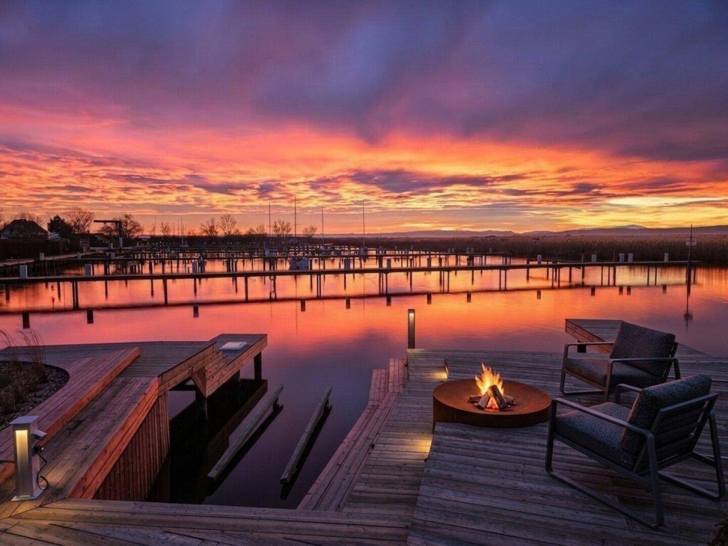 Gemütliche Holzterrasse mit Feuerstelle und atemberaubendem Blick auf den See bei Sonnenuntergang.