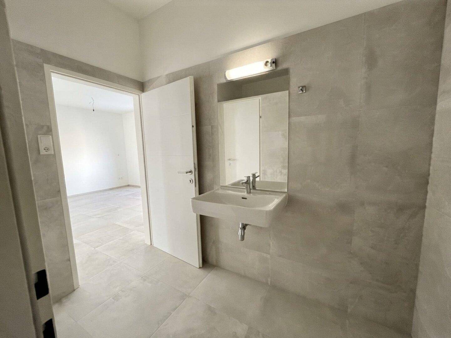 Modernes Badezimmer mit grauen Fliesen, wandmontiertem Waschbecken und Spiegel.