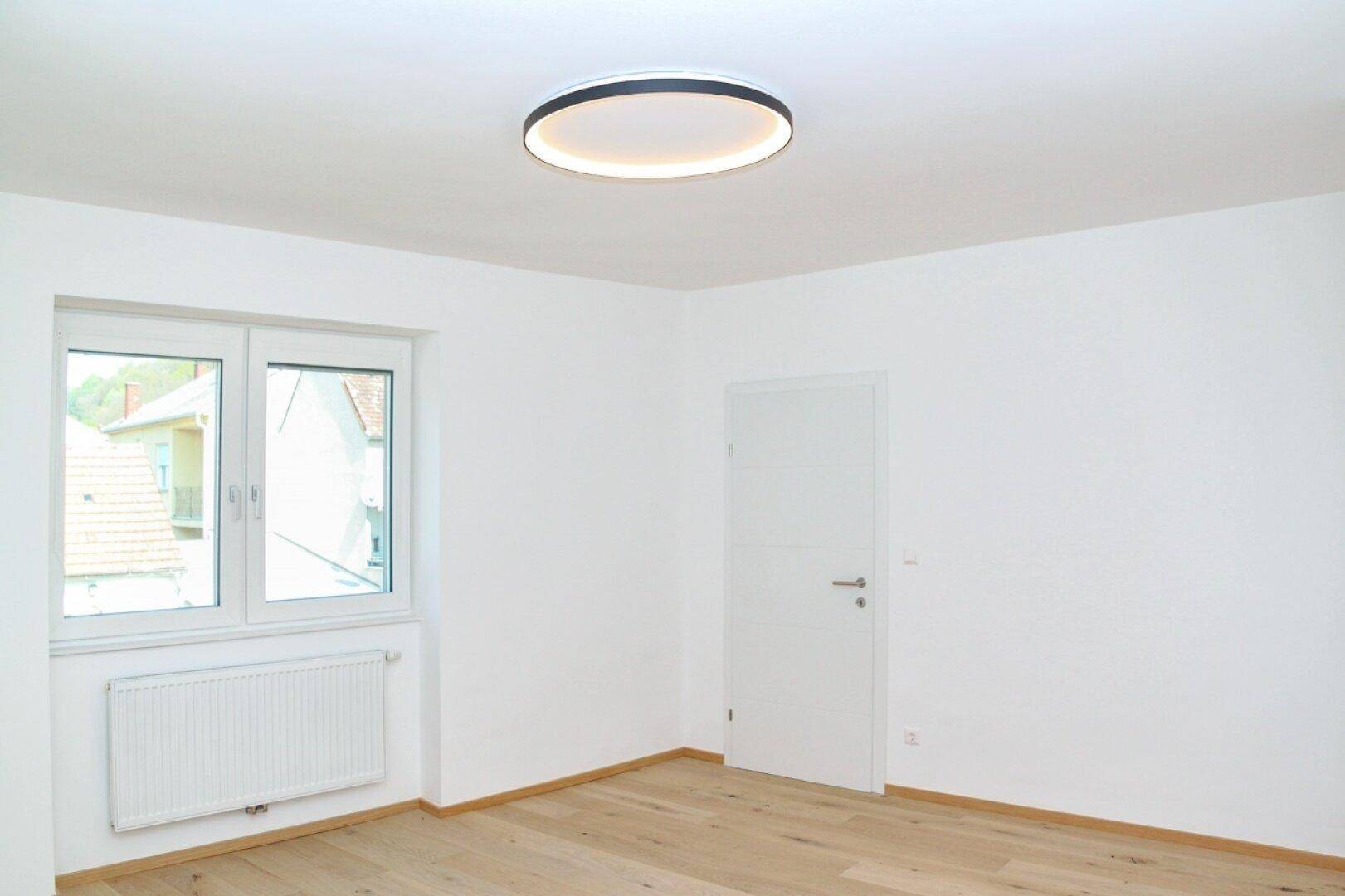 Geräumiger, heller Raum mit Laminatboden, großem Fenster und moderner Deckenbeleuchtung.