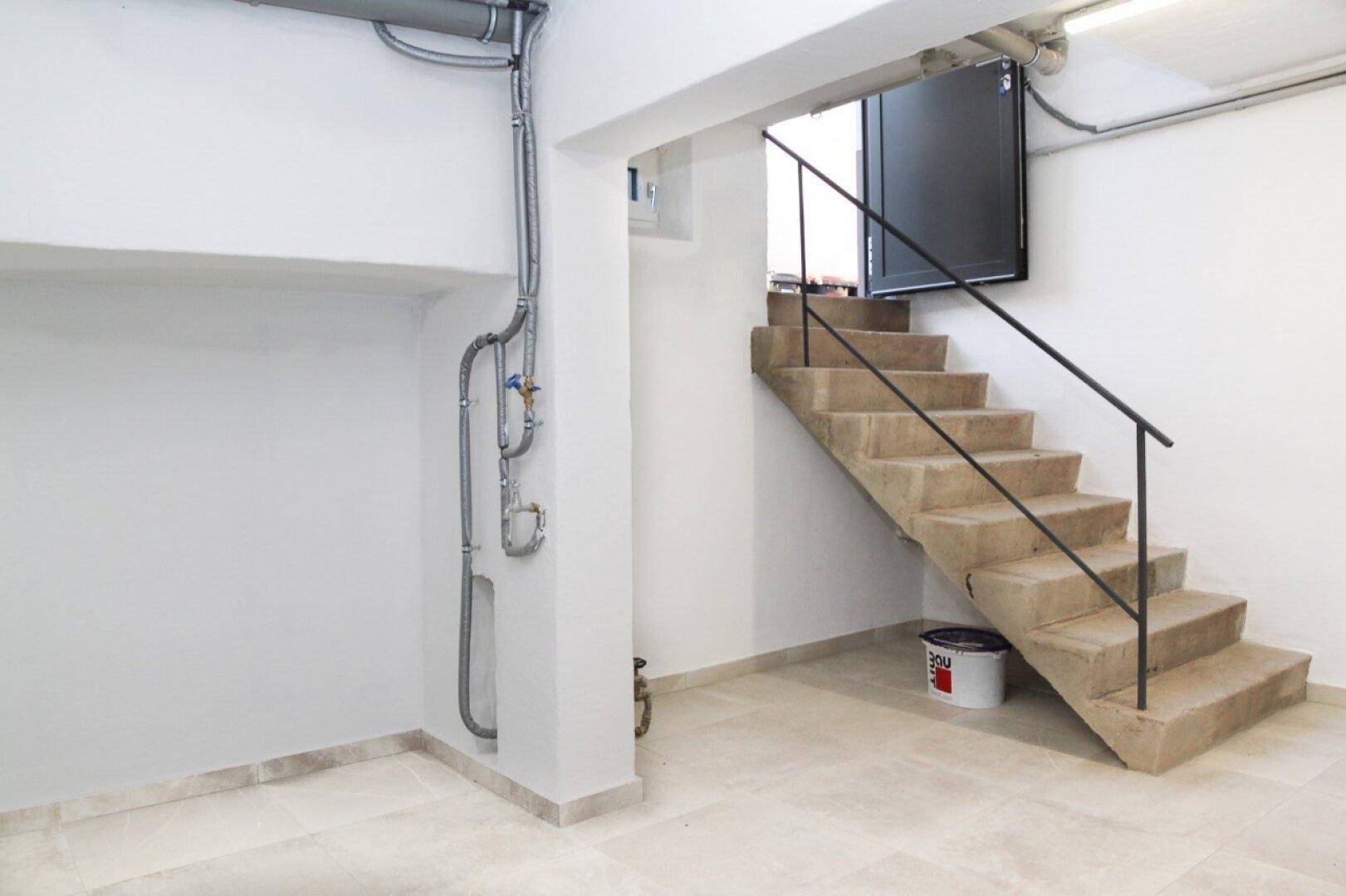 Kellerraum mit Betontreppe, sichtbaren Leitungen und Fliesenboden.