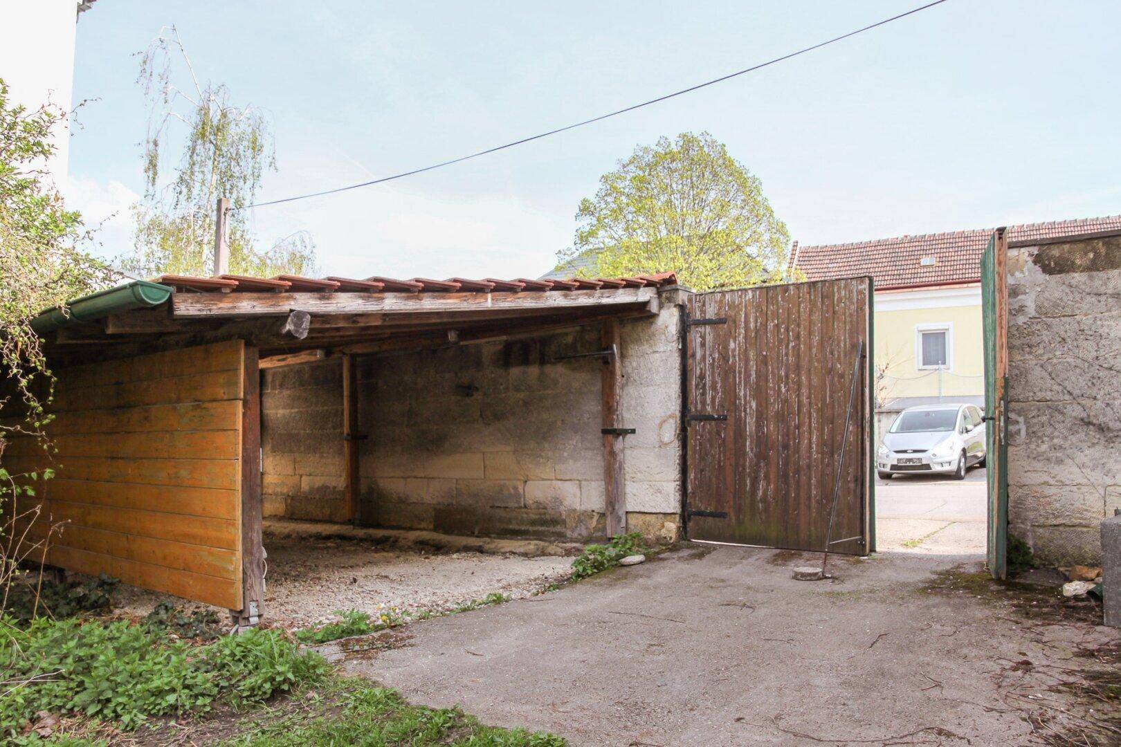 Einfacher Holzunterstand oder Carport mit Holztor und Schotterboden auf einem Grundstück.
