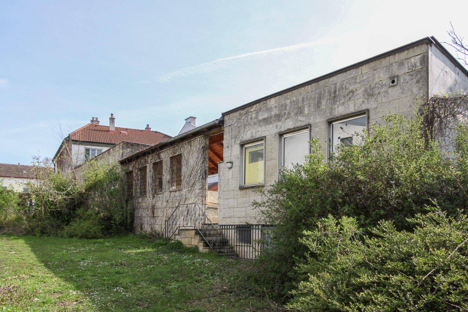 Langes Gebäude mit Steinfassade und überwachsenen Wänden, umgeben von einem ungepflegten Garten.