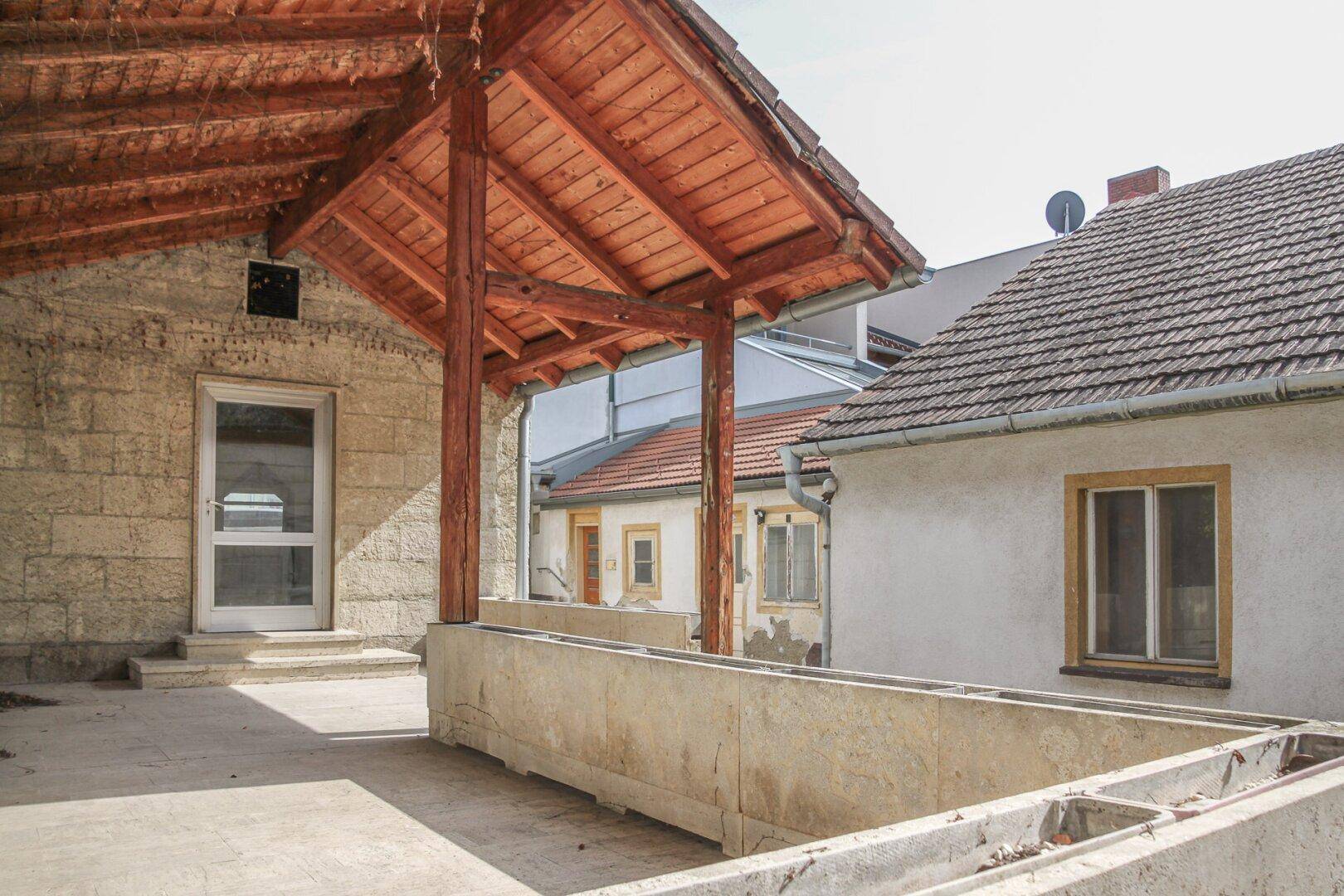 Überdachte Terrasse mit Holzbalken und Betonboden, angrenzend an ein älteres Gebäude.