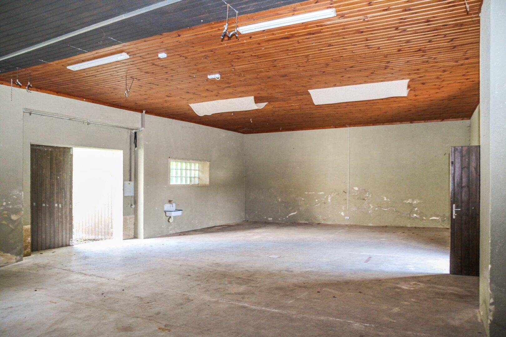 Geräumiger, leerer Raum mit Holzbalkendecke, Betonboden und einem kleinen Waschbecken an der Wand.