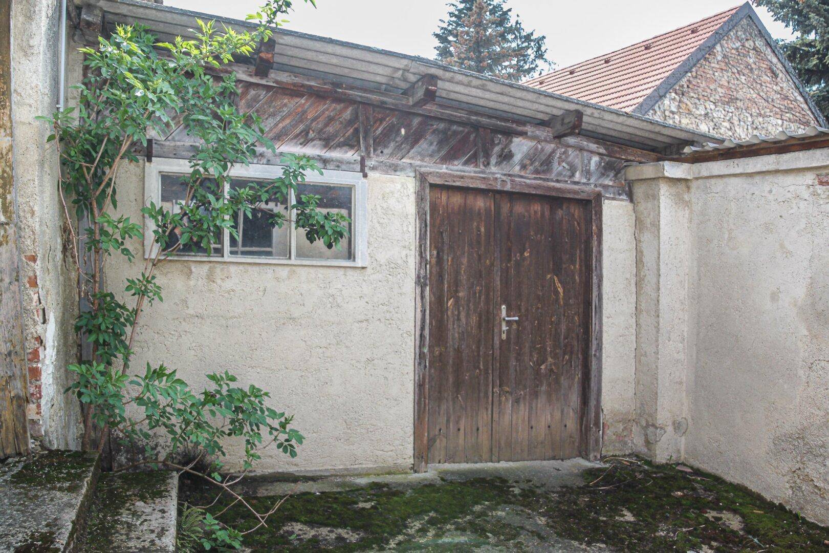 Kleiner Holzschuppen mit Holztür und Fenster, umgeben von einer verputzten Mauer und Grün.