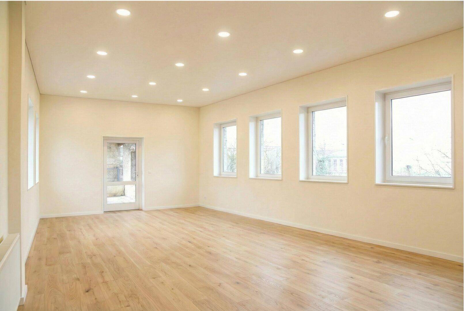 Heller, frisch renovierter Raum mit hellem Holzboden, vielen Einbaustrahlern und großen Fenstern.