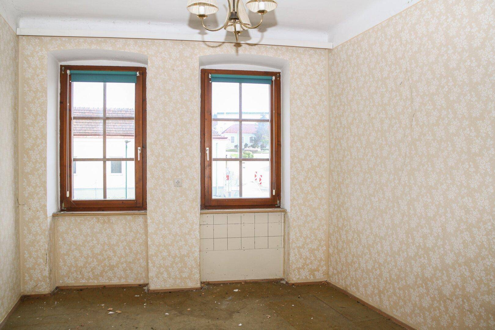 Leerer Raum mit alter gemusterter Tapete, zwei Fenstern und einem Kronleuchter an der Decke.