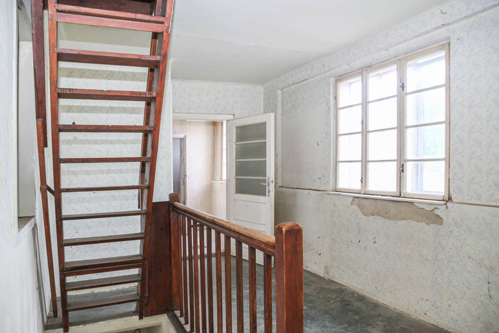Treppenhaus mit Holztreppe und Geländer, alter Tapete und einem Fenster, das Licht spendet.