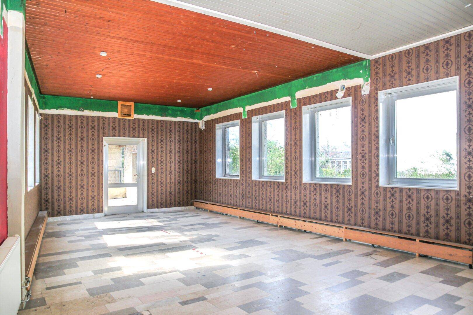 Großer Raum mit gemusterter Tapete, Holzdecke und mehreren Fenstern, renovierungsbedürftig.