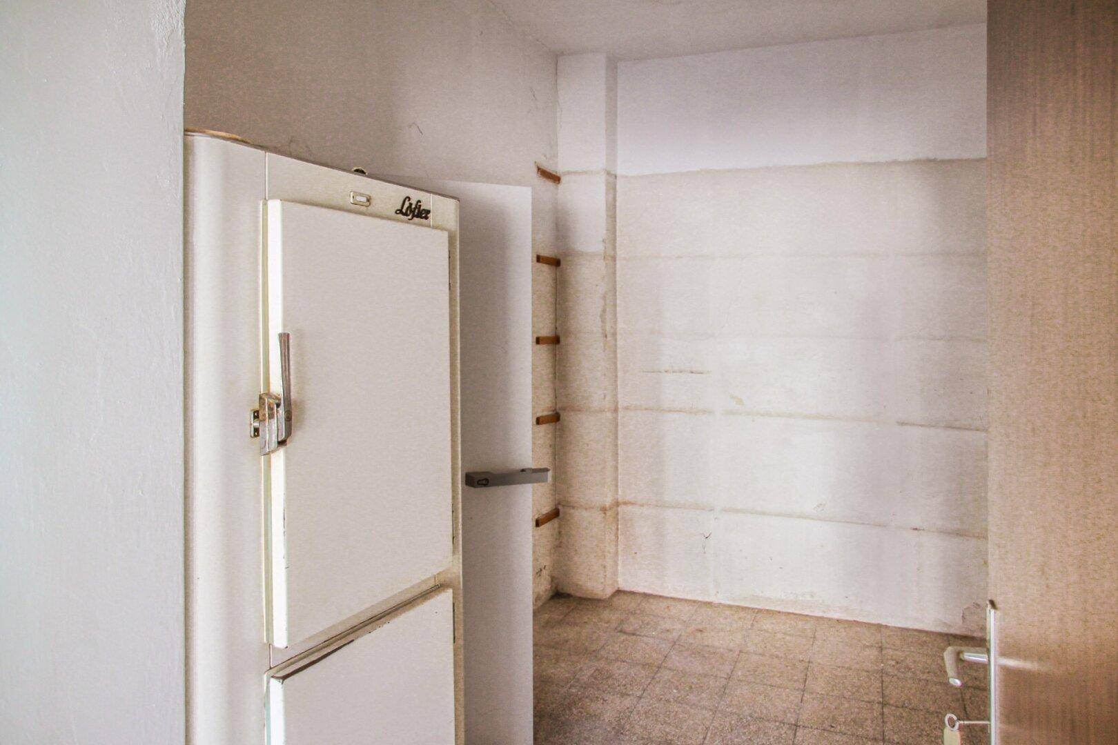 Leerer Raum mit altem Kühlschrank, gefliestem Boden und schlichten weißen Wänden.