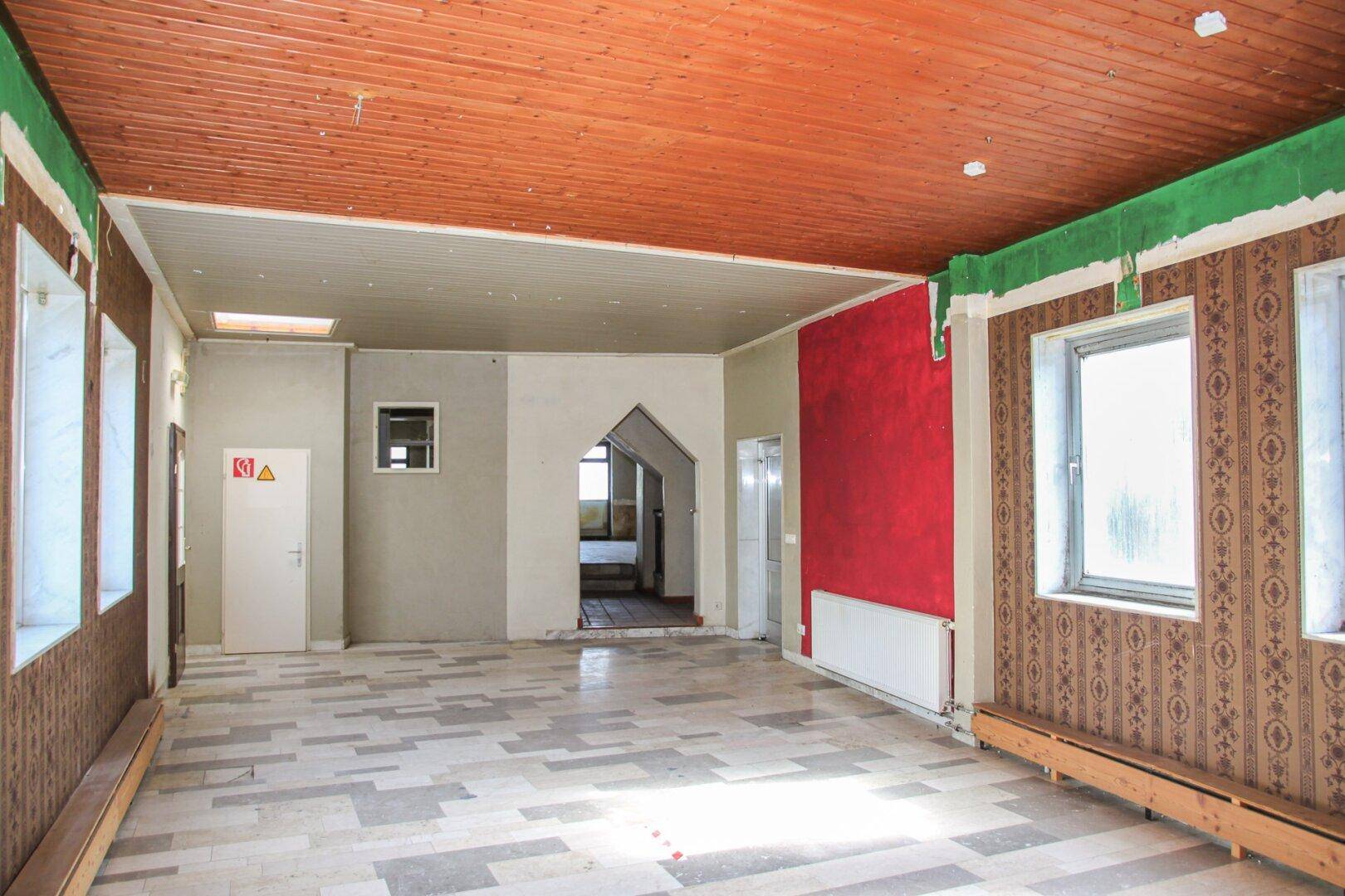 Geräumiger Raum mit Holzdecke, gemusterter Tapete und roter Akzentwand, renovierungsbedürftig.