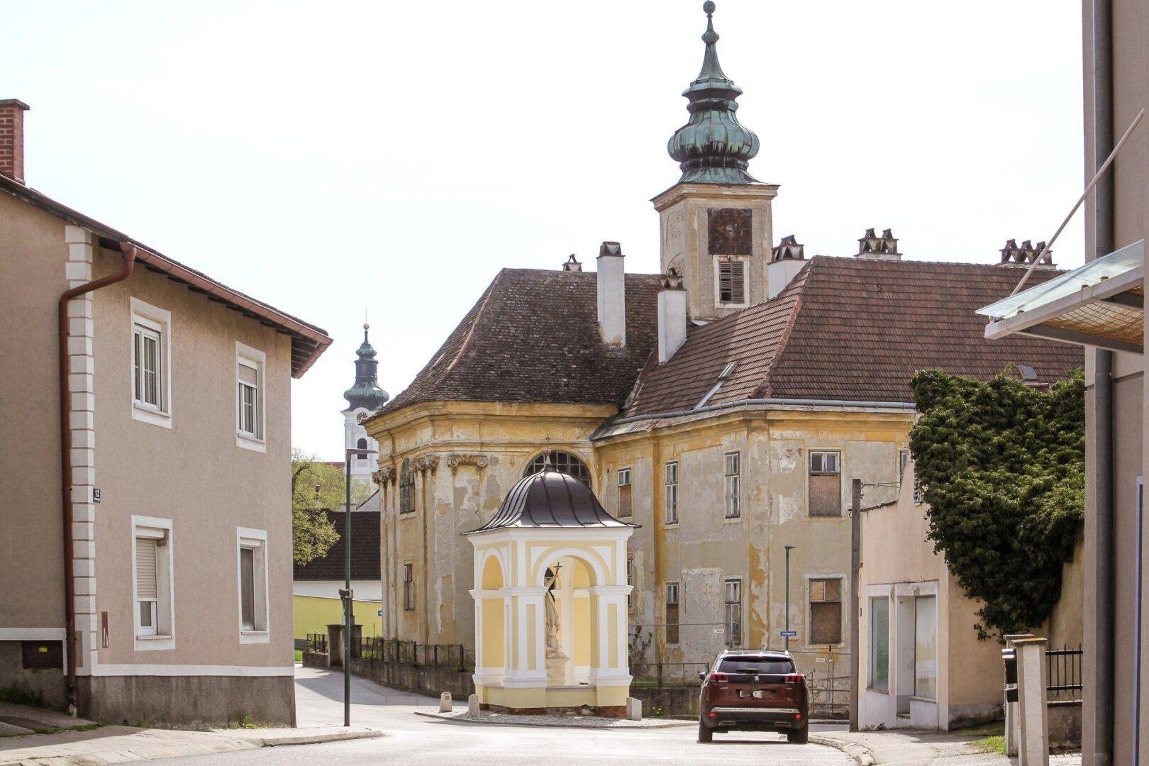 Historische Straße mit alten Gebäuden, einem Denkmal und einem geparkten Auto.