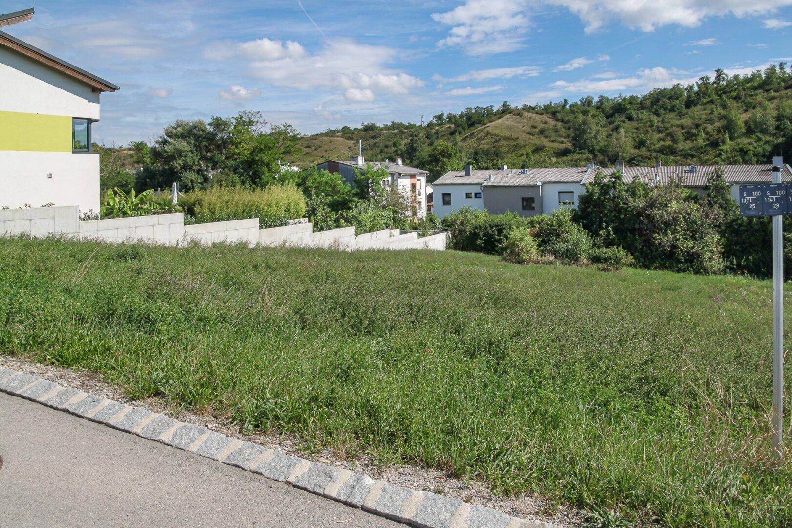 Unbebautes Grundstück mit hohem Gras, umgeben von modernen Häusern und grünen Hügeln.