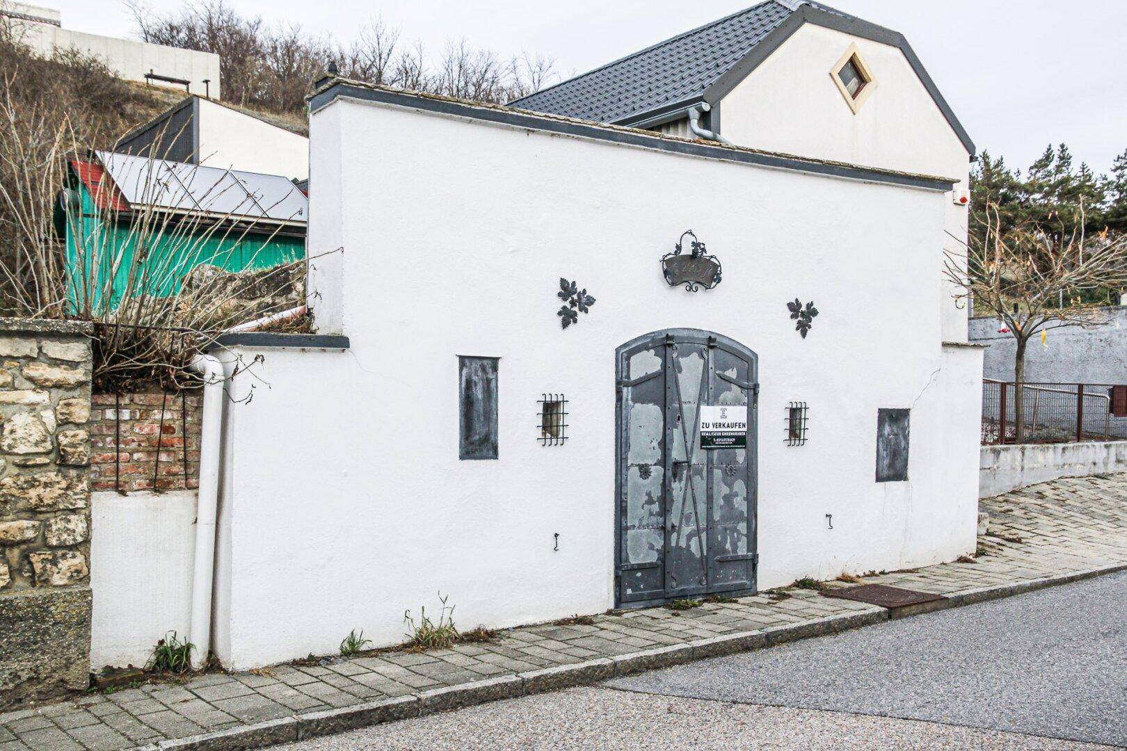 Weiß verputzte Fassade eines Gebäudes mit dunkler Eingangstür und kleinen Fenstern an einer Straße.
