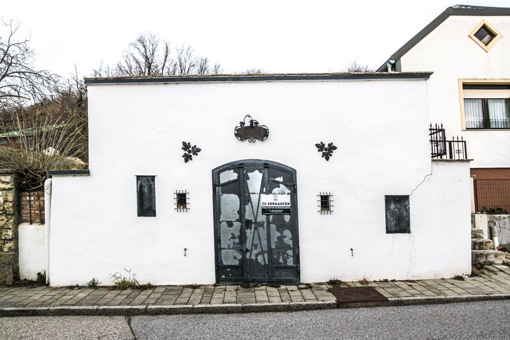 Ansicht der weißen Gebäudefassade mit rustikaler Tür und einem 'Zu Verkaufen'-Schild an der Straße.