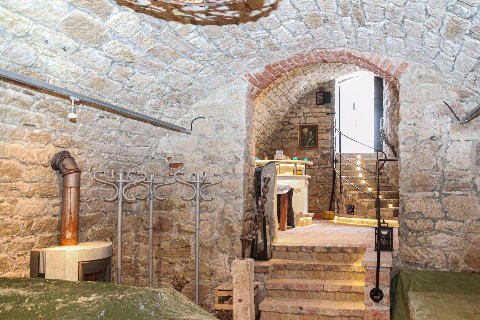 Gemütlicher Gewölbekeller mit Kaminofen, Garderobenständern und Zugang zur beleuchteten Steintreppe.