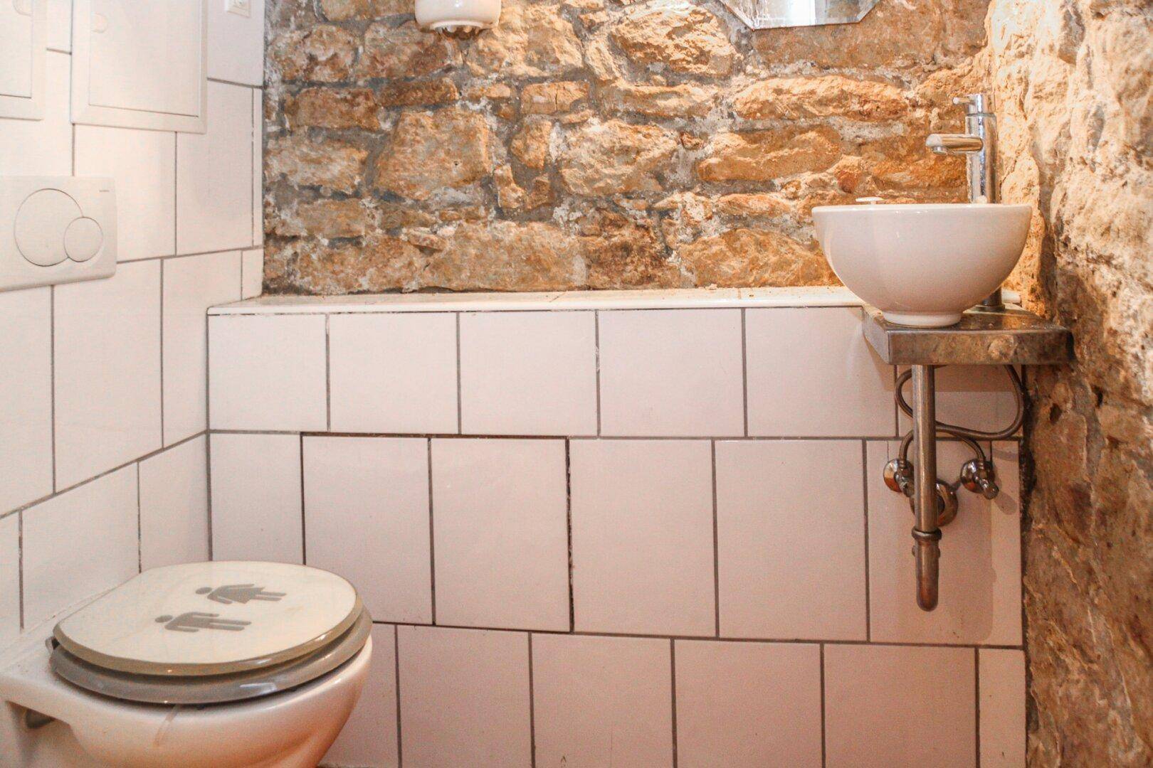 Kleine Gästetoilette mit weißen Fliesen, Wand-WC und einem runden Aufsatzwaschbecken auf Metallkonsole.