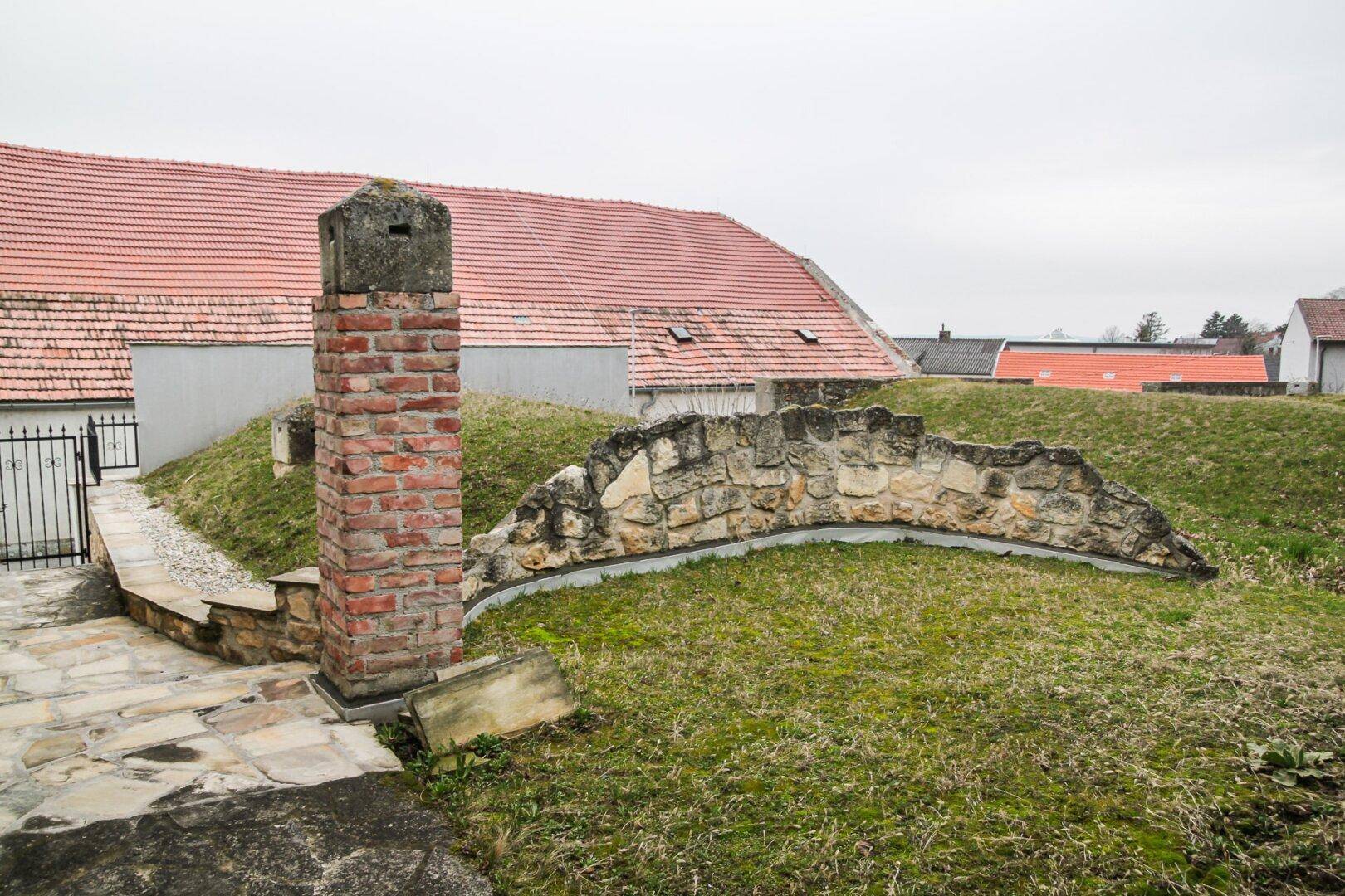 Gartenbereich mit Rasenfläche, einer Ziegelsteinsäule und einer geschwungenen Natursteinmauer im Vordergrund.