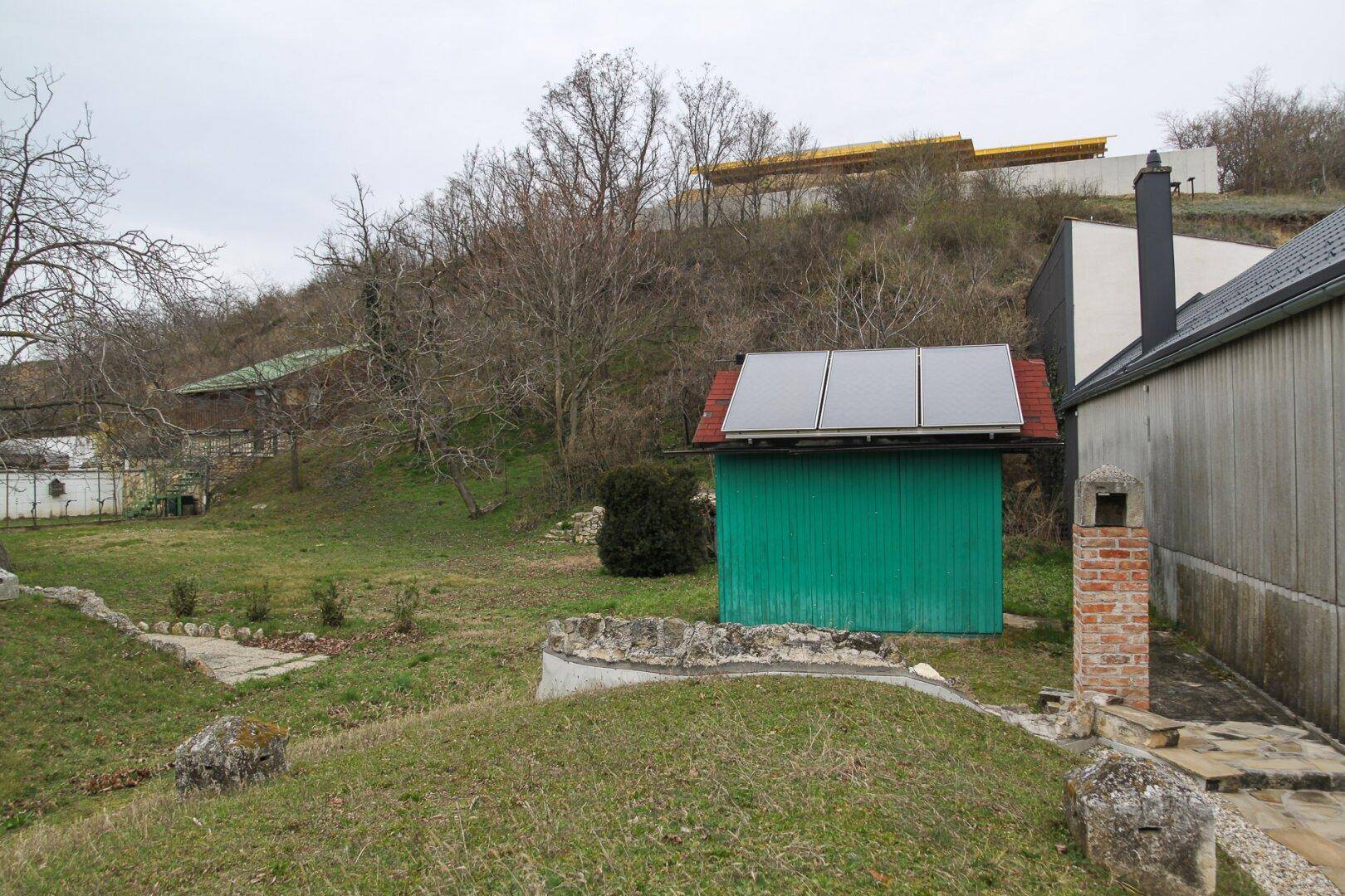 Garten mit grünem Schuppen, Solarpaneelen auf dem Dach und einer Rasenfläche mit alten Bäumen.