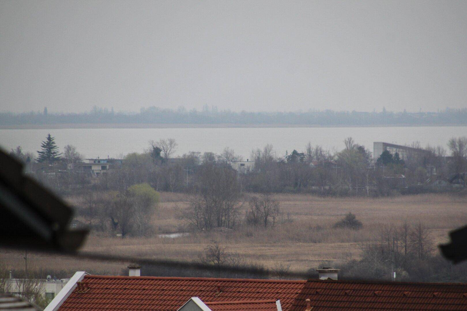 Weitläufiger Blick auf einen See oder Fluss über Dächer und Felder an einem bewölkten Tag.