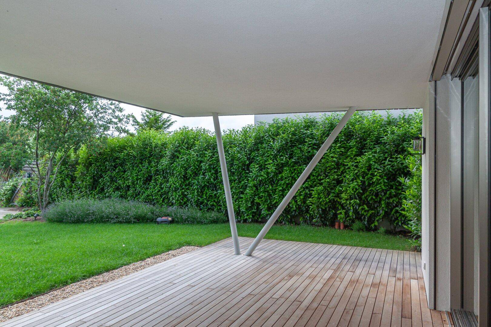 Überdachte Holzterrasse mit schrägen Stützen, umgeben von einer hohen grünen Hecke und Rasenfläche.