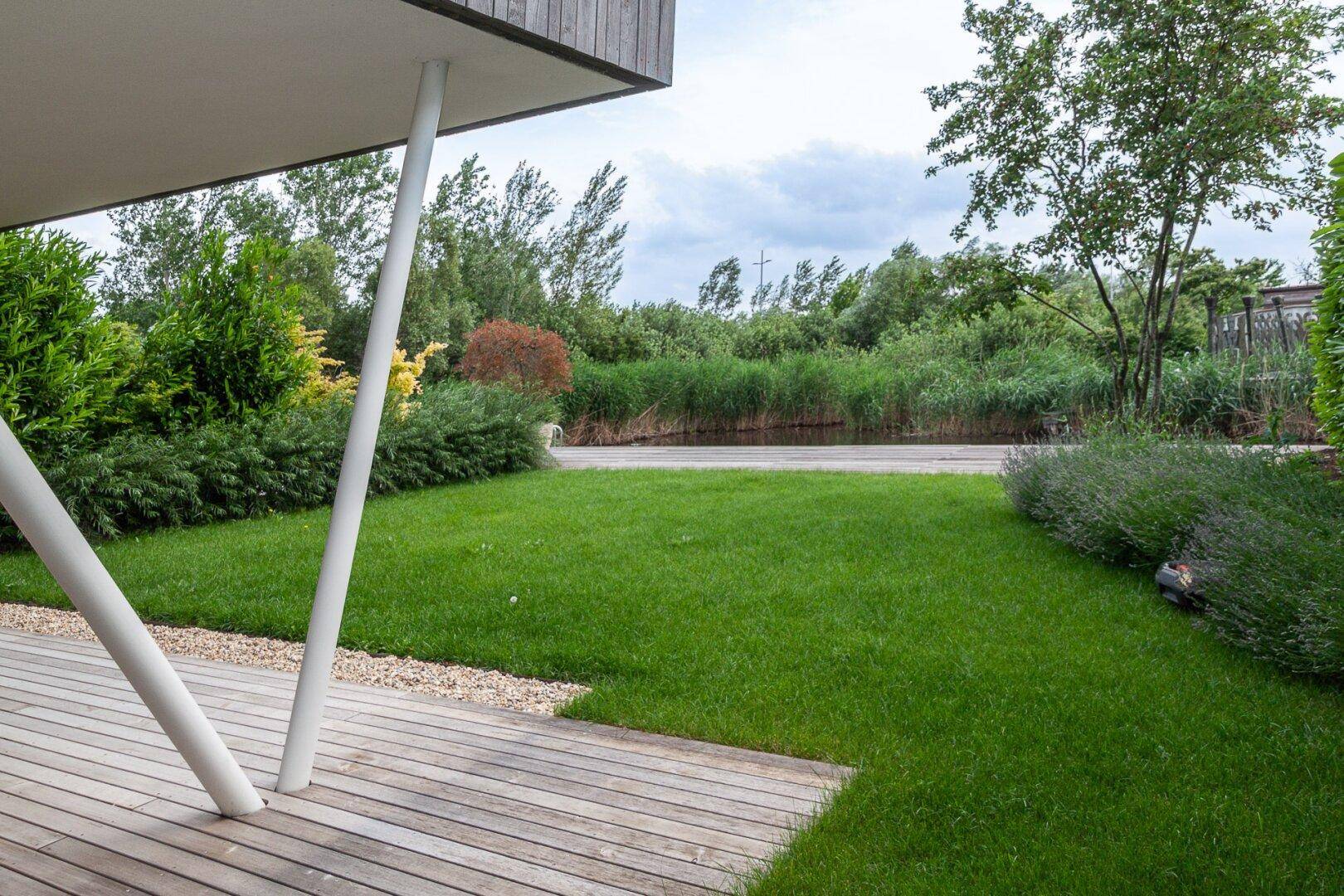 Blick von der überdachten Terrasse auf den gepflegten Garten mit Rasen und Schilf am Wasser.