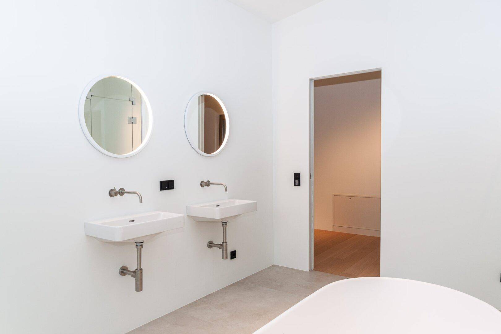 Modernes Badezimmer mit zwei Waschbecken und runden Spiegeln, offen zu einem angrenzenden Raum.
