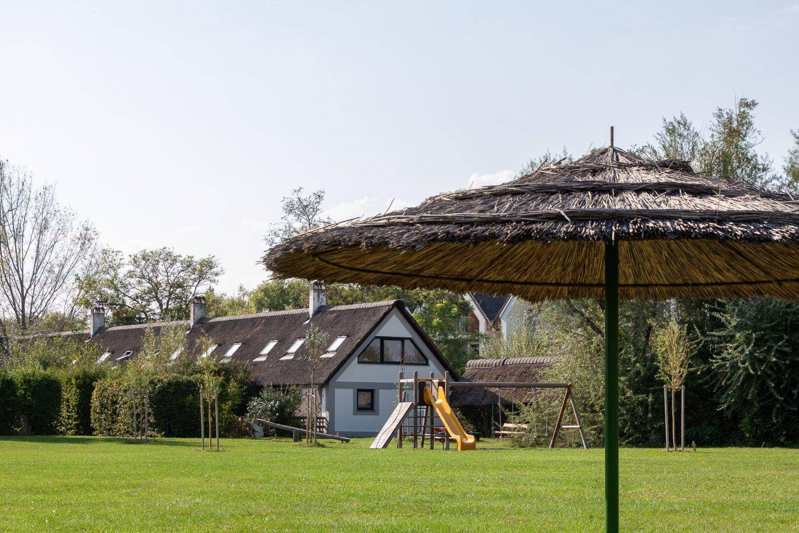 Weitläufiger Gartenbereich mit Spielplatz und Reetdachhäusern im Hintergrund.
