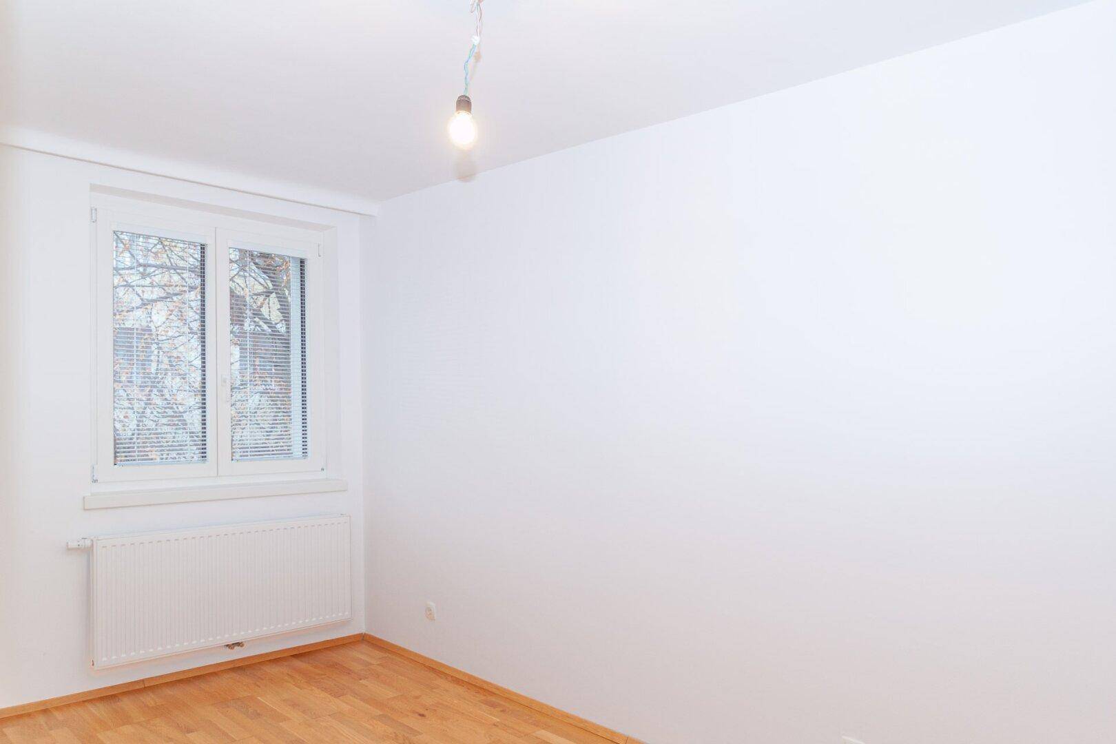 Helles, leeres Zimmer mit Holzfußboden, weiß gestrichenen Wänden und einem großen Fenster.