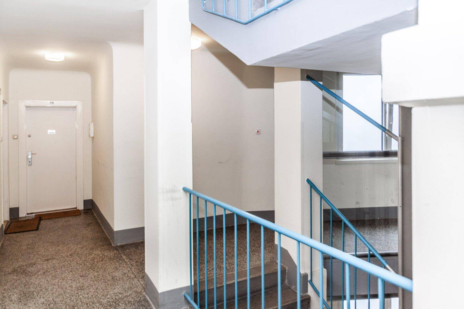 Treppenhaus und Flur eines Mehrfamilienhauses mit blauen Geländern und Terrazzoboden.