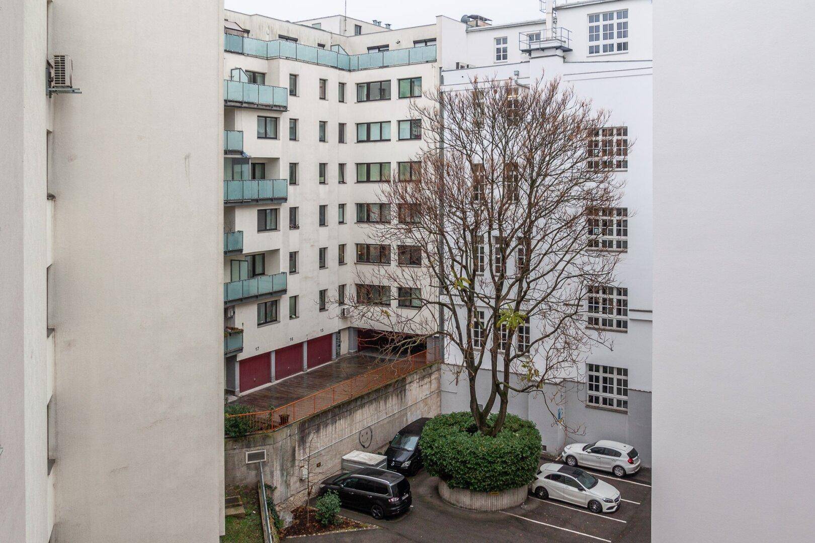 Innenhof mit Parkplätzen, einem Baum und den Fassaden der umliegenden Gebäude.
