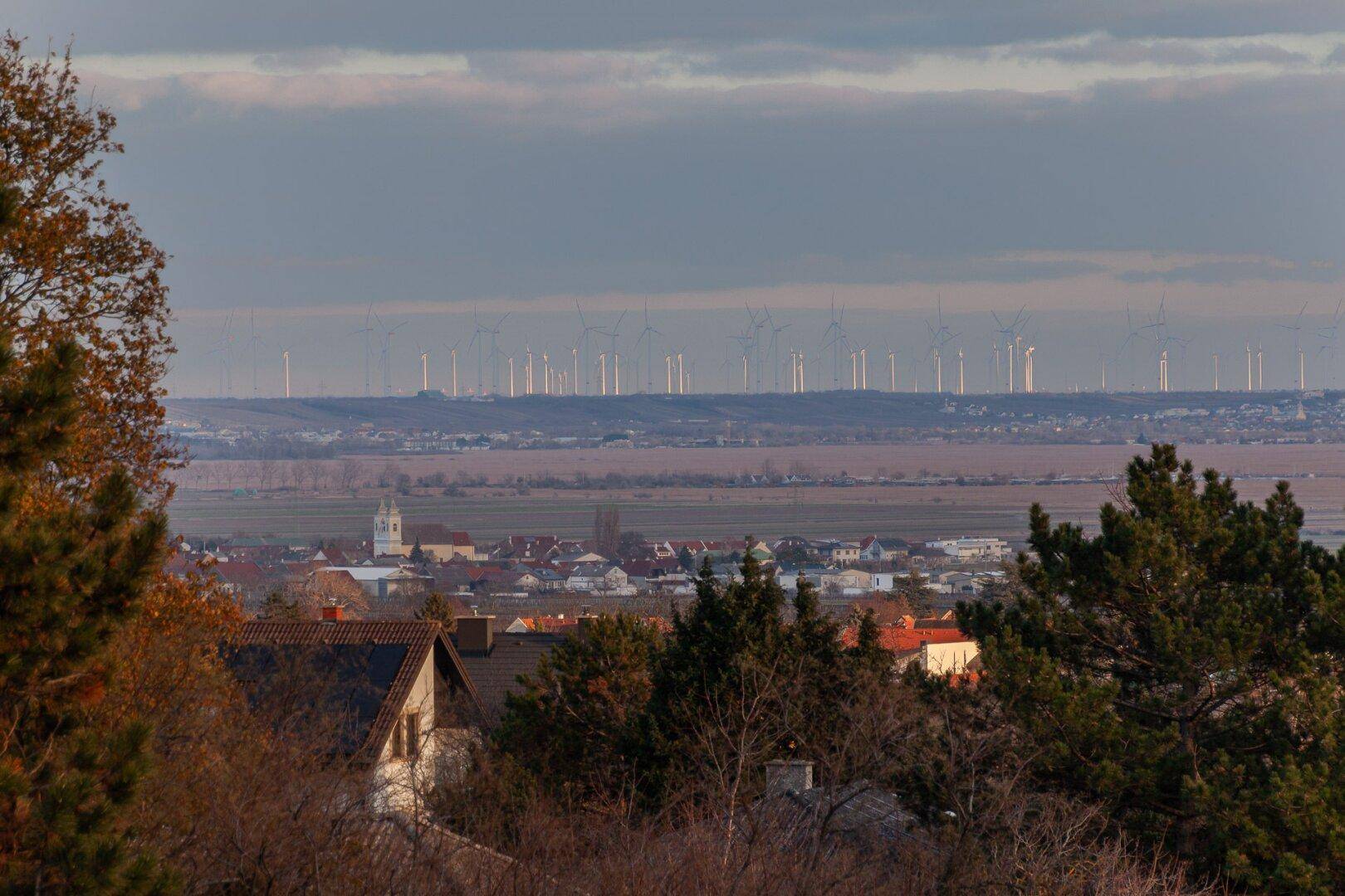 Weitläufiger Blick über die Landschaft mit Windrädern am Horizont und einem Dorf im Vordergrund.