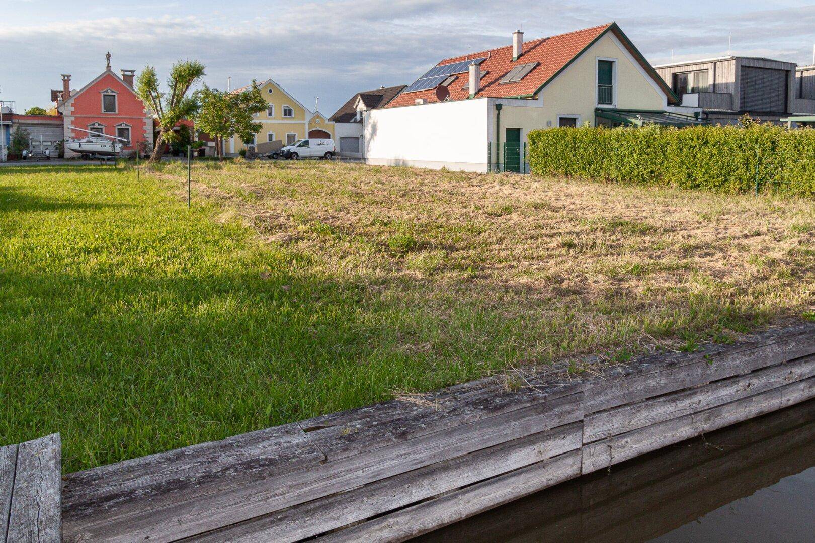 Grüne Rasenfläche am Kanalufer mit traditionellen Häusern und einem gepflegten Garten im Hintergrund.
