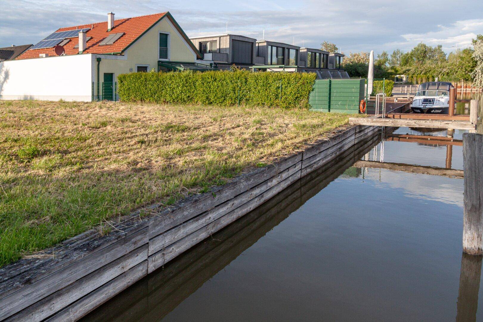 Uferbereich mit Holzverkleidung und Rasenfläche, dahinter ein Haus mit Solaranlage und Hecke.