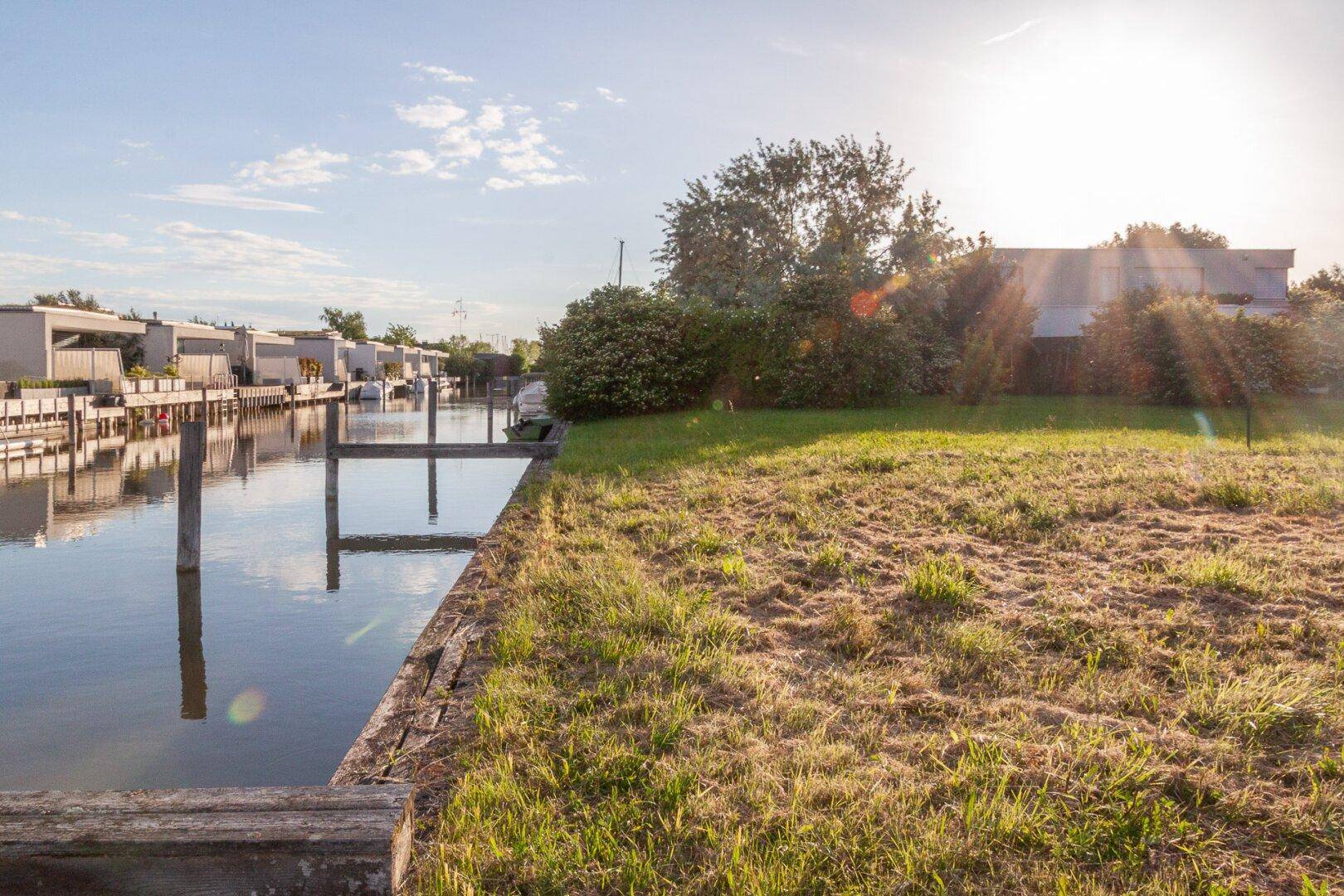 Sonnige Uferlandschaft mit Grasfläche und Holzpfählen am Wasser unter einem strahlend blauen Himmel.