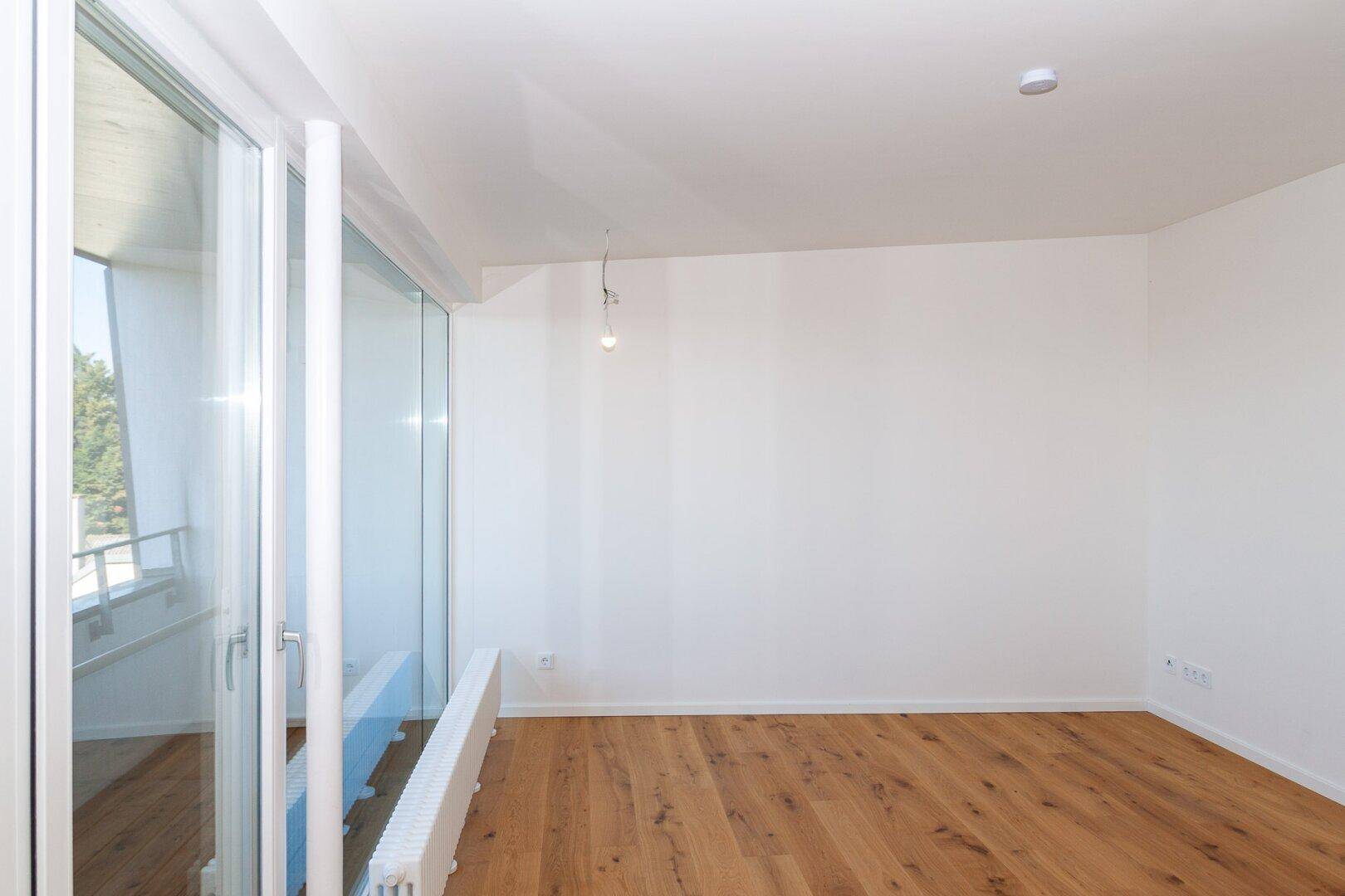 Leerer, heller Raum mit Holzboden und großen Fenstern, die Zugang zu einem Balkon bieten.
