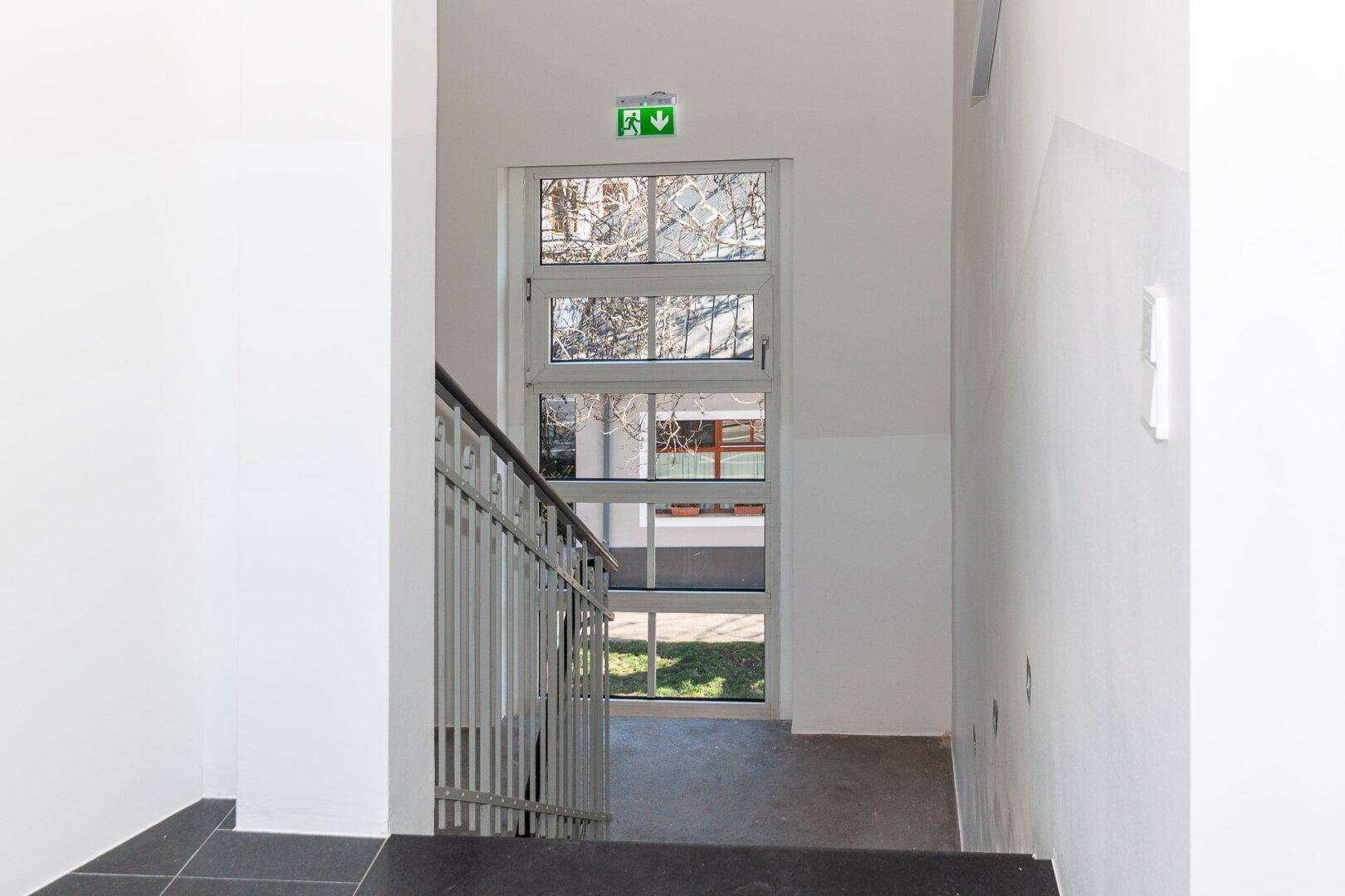 Helles Treppenhaus mit großen Fenstern und einem Notausgangsschild, das Tageslicht hereinlässt.