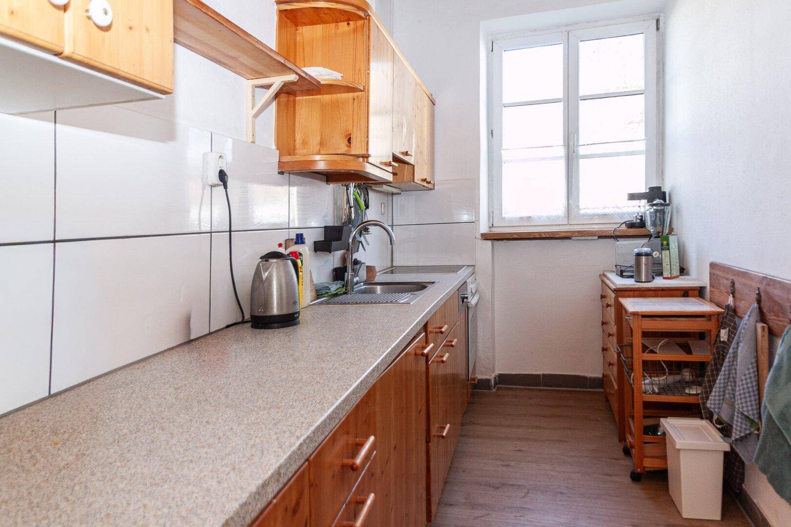 Funktionale Küche mit Holzschränken, heller Arbeitsplatte und Fenster für Tageslicht.