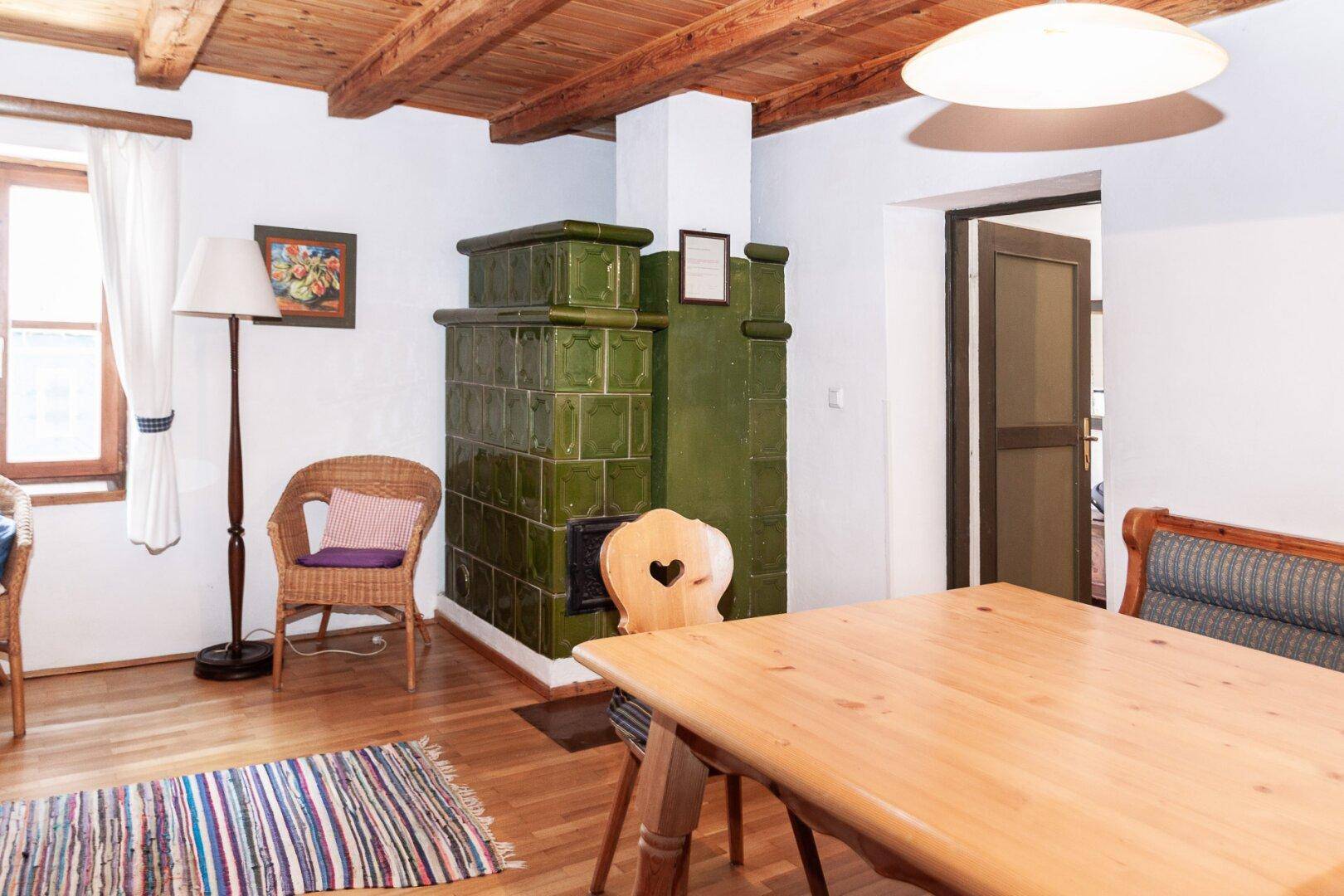 Rustikaler Essbereich mit großem Holztisch, grünem Kachelofen und Holzbalkendecke.
