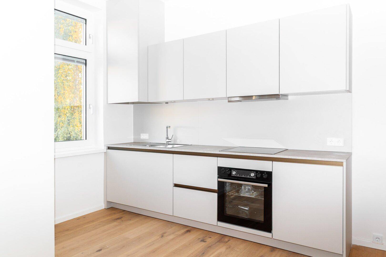 Helle Küche mit modernen weißen Schränken, Holzfußboden und Fensterblick ins Grüne.